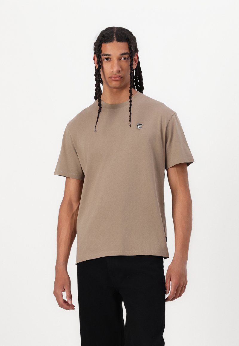 Brun bomulds T-shirt med korte ærmer, rund hals og et lille logo på brystet. Kombination med sorte bukser. Glat tekstur, afslappet pasform.