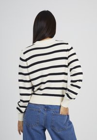 Donna con lunghi capelli scuri, che indossa un maglione bianco con strisce nere orizzontali e jeans blu, in posa con una mano nella tasca posteriore.