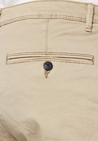 Beige cotton pants feature a back pocket with a horizontal opening and a black button. The fabric is smooth with a slight sheen.