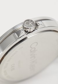 Montre en acier inoxydable argenté avec un cadran rond et une couronne saillante arborant le logo "CK". Accents texturés sur le côté.