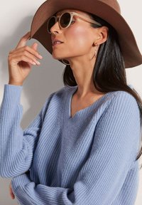 Hellblau gestrickter Pullover mit V-Ausschnitt, langen Ärmeln und strukturiertem Stoff. Begleitet von einem breitkrempigen braunen Hut und runden Sonnenbrillen.