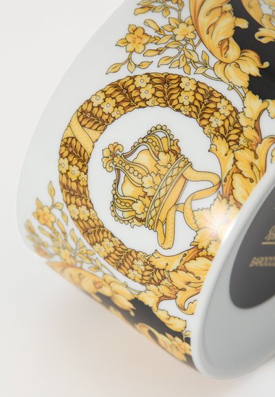 Λευκό κεραμικό φλιτζάνι με περίπλοκα χρυσά λουλουδένια σχέδια, μοτίβο κορώνας και μαύρη λεπτομέρεια κατά μήκος της βάσης. Διακρίνονται υφές και λεπτομέρειες.