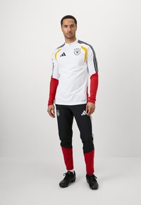 Λευκή μακριά μπλούζα με κόκκινες μανσέτες, κίτρινες και μαύρες λεπτομέρειες και έμβλημα. Μαύρο παντελόνι με κόκκινες κατώτερες περιοχές και λογότυπο Adidas. Μαύρα παπούτσια.