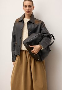 Czarna skórzana torba typu tote o zaokrąglonym kształcie, z krótkimi uchwytami i srebrnymi ćwiekami na dnie, w połączeniu z szarym żakietem i kremowym swetrem na brązowej spódnicy.