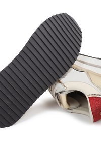 Zapatilla con suela de goma negra que presenta ranuras horizontales, parte superior sintética en blanco, detalles de purpurina dorada y roja, y un diseño aerodinámico.