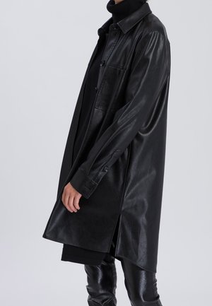 Persoon met een lange zwarte leren overhemdjas over een zwarte coltrui en zwarte broek, van opzij gefotografeerd tegen een grijze achtergrond.