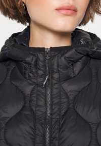 Giacca nera trapuntata con cappuccio, dotata di una cerniera argentata sul davanti e una texture liscia e lucente con una leggera brillantezza.