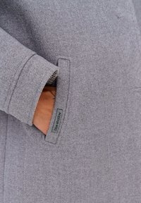 Manteau en laine gris avec un design structuré, doté d'une poche avant et d'une doublure à carreaux subtile. Étiquette "Blue Shadow" visible.