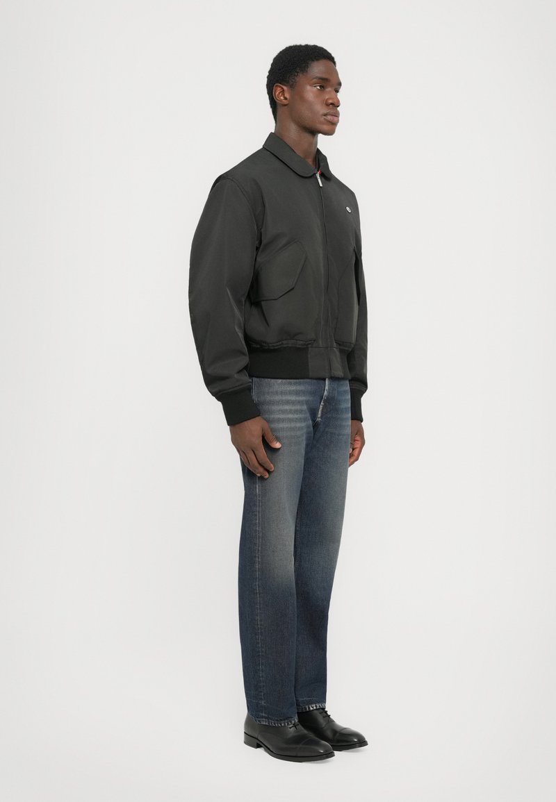 Veste bomber noire avec deux grandes poches avant, poignets et col côtelés, associée à un jean bleu droit et des chaussures noires.