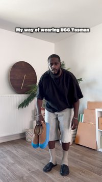 Pantofle UGG Tasman s hnědým svrškem a modrou gumovou podrážkou drží v ruce. Člověk má na sobě černé tričko, šedé šortky a béžové ponožky, stojí uvnitř.