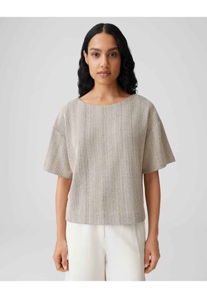 Femme aux longs cheveux noirs portant un haut à manches courtes à rayures verticales beige et noires et un pantalon blanc, debout devant un fond uni.