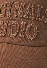 Brun cap med struktureret stof, prydet med slidte broderier af ordene "Originals" og "Audio" i brun tråd, der viser det underliggende materiale.
