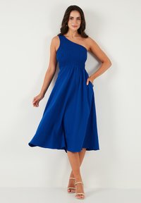 One-shoulder, blue dress with smocked bodice, flowing skirt, and side pockets. Made of lightweight fabric, featuring a natural texture.