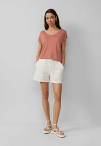 Rozā V-veida t-krekls, īsas baltas bikses ar aukliņu, un baltas siksnainas čības. Gluda auduma, brīvs piegriezums, ar redzamām kabatām biksēs.