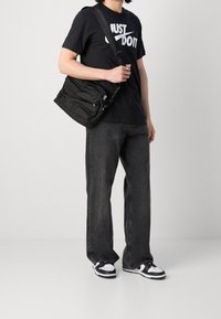 Person wearing black "Just Do It" T-shirt, gray jeans, black-and-white sneakers, and carrying a black shoulder bag with multiple zip compartments.