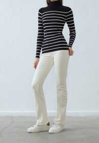 Suéter de cuello alto a rayas en blanco y negro con textura acanalada, combinado con pantalones acampanados en crema y zapatillas blancas sobre un suelo liso.