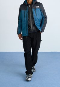 Padded jacket w kolorze zielono-niebieskim i czarnym, z kapturem i kieszeniami na zamek. Nosi się go na czarnej skórzanej kurtce i w zestawieniu z czarnymi spodniami.