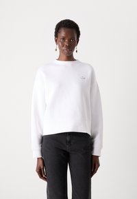 White sweatshirt with a round neckline and cropped design, featuring a small embroidered logo on the chest. Paired with dark jeans.