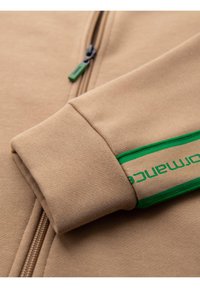Tessuto di felpa beige con chiusura a zip, dotato di un accento logo verde sul polsino della manica e superficie testurizzata.