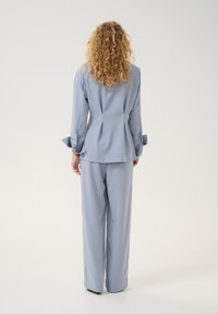 Abito su misura azzurro chiaro con maniche lunghe, dettagli plissettati sul retro e accenti sui polsini. I pantaloni a gamba dritta completano il look. Texture del tessuto liscia.
