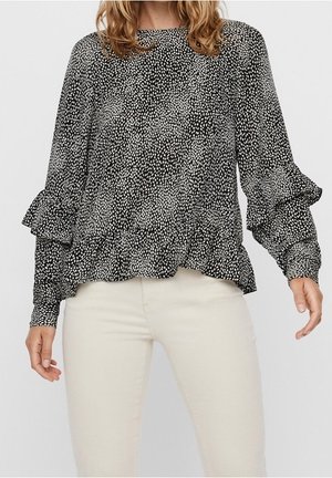 Mujer con blusa de manga larga con estampado blanco y negro, con puños y bajo fruncidos, combinada con pantalones beige claros.