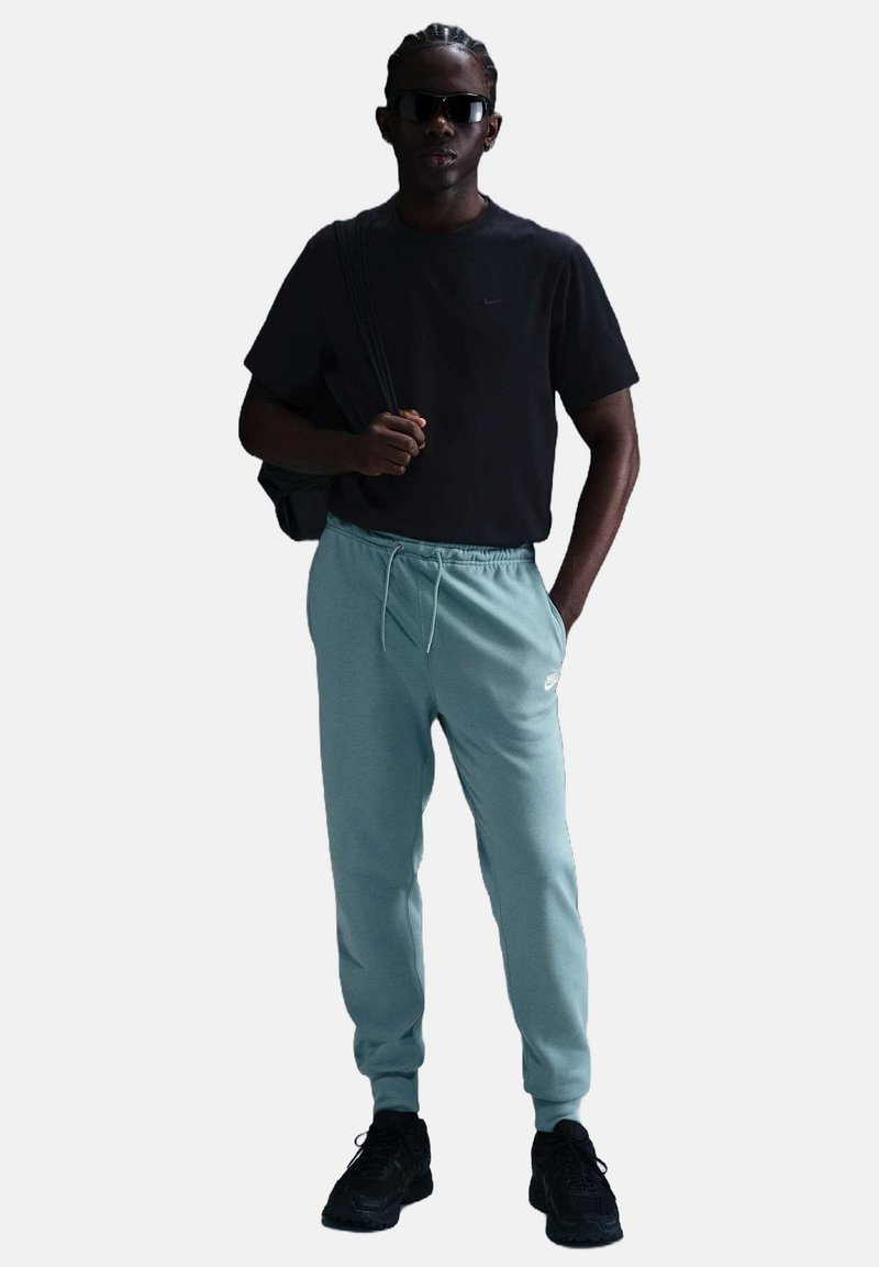 Camiseta negra, pantalones de sudadera azul claro y zapatillas negras. Los accesorios incluyen gafas de sol y una bolsa negra. Atuendo atlético casual.