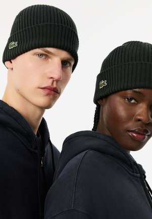 Los gorros de punto acanalado negros presentan un pequeño logotipo en verde y rojo. Ambos son usados por individuos en sudaderas oscuras con vistas en primer plano de sus rostros.