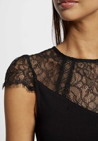 Vestido negro con mangas cortas de encaje transparente y patrones florales intrincados, y un torso liso y sólido. Detalles de encaje texturizado en el escote.