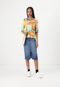 Bunten Kurzarm-Oberteil mit abstrakten Mustern in Orange, Türkis und Weiß; kombiniert mit einem knielangen Jeansrock und weißen Sneakers.