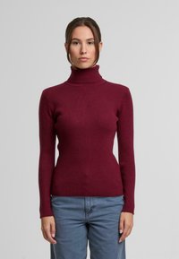 Vínová žebrovaná roláková svetr s dlouhými rukávy, vypasovaným střihem a texturovanou tkaninou, v kombinaci s světle modrými kalhotami volného střihu.