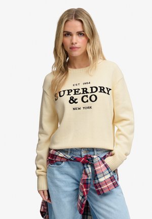 Lyshåret kvinde iført en cremefarvet Superdry sweater, lyseblå jeans og en rød og blåternet skjorte bundet om taljen.