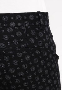 Černé kalhoty se vzorem, které obsahují jemný design kruhových motivů a malých hvězdiček, s jednou zadní kapsou a poutky na opasek. Měkká textilie.