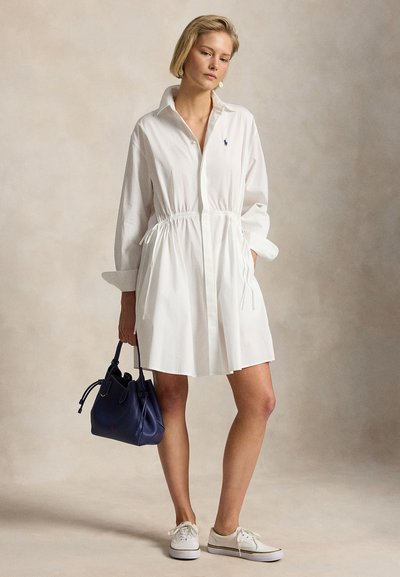 Kvinne iført hvit langermet kjole med snøring i midjen, hvite joggesko, og holder en mørkeblå håndveske foran en beige bakgrunn.