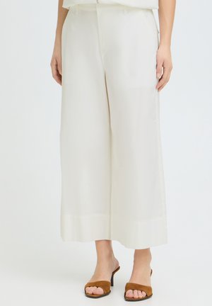 Kvinde iført hvide, vide, korte bukser og brune åbne hælede sandaler, stående mod en ensfarvet baggrund.