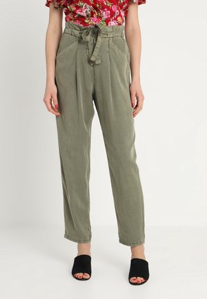 Kvinde iført olivengrønne bukser med høj talje og bundet bælte, en rød blomstret top og sorte mulehøjehælede sko med åbne tæer, stående mod en ensfarvet baggrund.