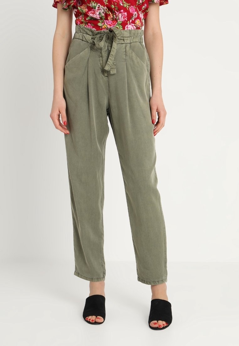 Femme portant un pantalon taille haute vert olive avec une ceinture nouée, un haut rouge à motifs floraux, et des mules à talons ouvertes noires, debout devant un fond uni.