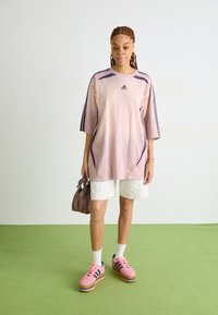 Prevelik svetlo roza športni jelovnik s vijoličnimi poudarki, v kombinaciji z belimi kratkimi hlačami s črtami in roza platformskimi supergami z modrimi črtami.