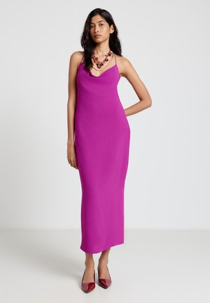 Donna in piedi con un maxi abito senza maniche color magenta con scollo drappeggiato, che indossa tacchi rossi e una collana rossa vistosa su uno sfondo semplice.