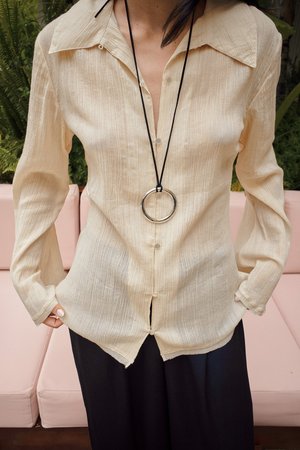 Camisa beige de botonadura con textura, cuello puntiagudo, mangas largas y dobladillo curvo. Llevada con un collar de péndulo negro.