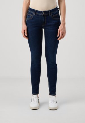 Person trägt dunkelblaue Skinny-Jeans und weiße Sneaker und steht vor einem schlichten hellen Hintergrund.