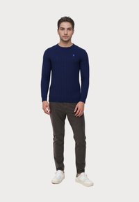 Jonge man die staat, gekleed in een knapblauwe ribtrui, donkergrijze broek en witte sneakers, tegen een effen lichte achtergrond.