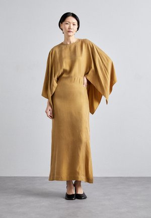 Robe longue couleur moutarde avec une silhouette ample et décontractée, dotée de manches chauve-souris et d'une texture lisse. Associée à des talons noirs.