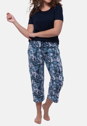 Vrouw draagt een zwart T-shirt met korte mouwen en blauwe korte broek met een wit en zwart bloemmotief, staat op blote voeten met gebogen armen.