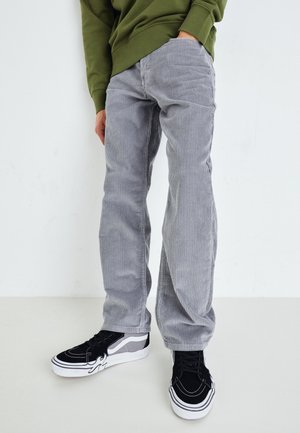 Personne portant un pantalon en velours côtelé gris clair, des baskets noires et blanches, et un sweat-shirt vert olive, debout sur un fond blanc.