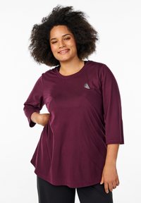 Bordeauxfarbene Langarm-Bluse mit rundem Ausschnitt und Dreiviertelärmeln, aus glattem Stoff gefertigt. Mit einem kleinen geometrischen Logo auf der Brust.