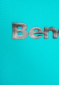 Tirkizna tkanina s riječju "Benetton" u uzdignutom srebrnom slovu, ističući glatku teksturu i kontrastne boje.