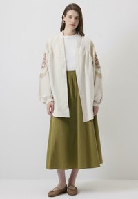 Jachetă lejeră de un bej deschis, cu broderie florală pe mâneci, purtată cu o fustă lungă de un verde măsliniu și balerini texturați de culoare maronie.