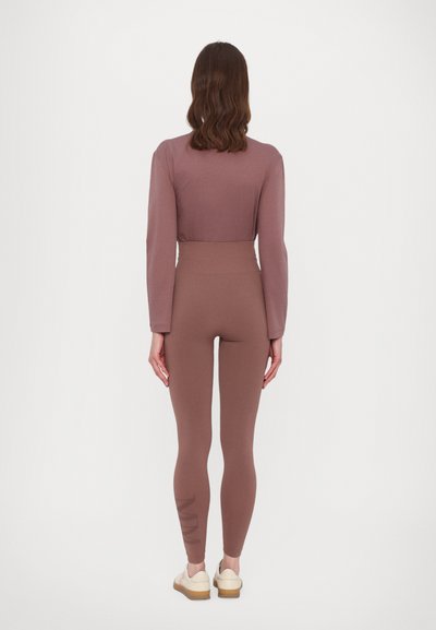 Brauner langärmeliger Oberteil kombiniert mit passenden hochgeschnittenen Leggings, die ein dezentes Logo am linken Bein aufweisen, vor einem einfarbigen Hintergrund.