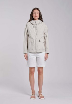 Kobieta w jasnobeżowej kurtce z kapturem, białych krótkich spodenkach do kolan i białych sandałach, stojąca na jednolitym szarym tle.