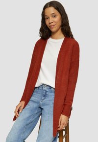 Cardigan couleur rouille avec un tissu texturé, à design ouvert à l'avant et manches longues, porté par-dessus une chemise blanche et associé à un jean bleu.